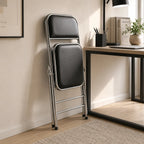 Shai Folding Chair folded beside a modern desk, showing its compact space-saving design and chrome frame.