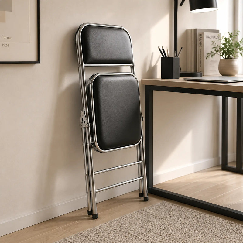 Shai Folding Chair folded beside a modern desk, showing its compact space-saving design and chrome frame.